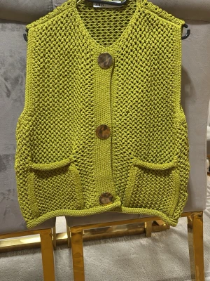 Stickad limegrön väst med stora knappar - Säljer en stickad limegrön väst med tre stora bruna knappar framtill och två praktiska fickor. Västen har en grovstickad struktur och är ärmlös med rundad hals. Perfekt för att addera färg till din outfit.