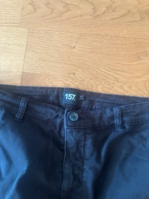 Svarta chinos från 157. - Säljer ett par svarta chinos från 157. De har klassisk passform med raka ben, fickor på sidorna och en knappstängning framtill. Materialet är mjukt och bekvämt, perfekt för dig som vill ha en stilren look. Passar dig som gillar enkel och tidlös stil.