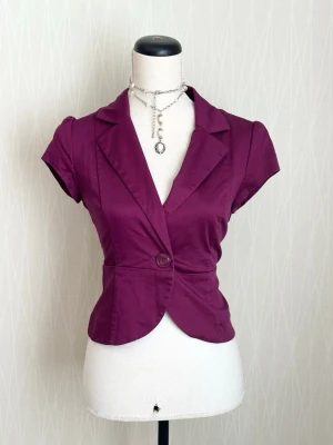 Kortärmad lila kavaj med knapp - Snygg figursydd kavaj i lila med korta ärmar och en enkel knapp framtill. Klassisk krage och djup v-ringning ger en modern look. Perfekt att styla över en topp eller klänning för en trendig outfit.