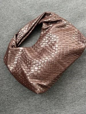 Brun handväska i croco-imitation - Säljer en brun handväska i croco-imitation med mjuk, rundad form och bred axelrem. Väskan har ett stilrent och trendigt utseende med struktur i materialet som påminner om krokodilskinn. Perfekt accessoar för att lyfta din outfit.