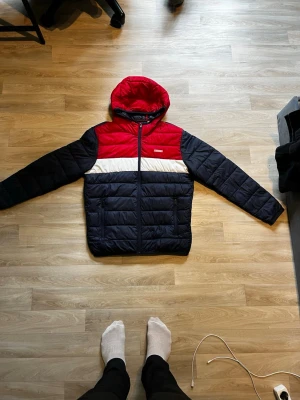 Jack & Jones pufferjacka röd/blå. Vi kan förhandla  - Snygg pufferjacka från Jack & Jones i färgblock med rött upptill, vitt band över bröstet och mörkblå nederdel och ärmar. Jackan har huva, dragkedja och quiltad design. Perfekt för kalla dagar och riktigt skön att bära.