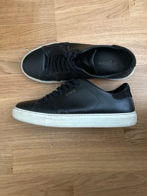 Svarta sneakers Axel Arigato läder - Stilrena svarta sneakers från Axel Arigato i slätt läder med svart mockadetalj vid hälen. Klassisk låg modell med vita gummisulor och svarta skosnören. Diskret guldfärgad logga på sidan. Perfekta för en clean och modern look.