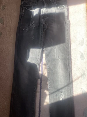 Svarta slitna skinny jeans - Säljer ett par svarta skinny jeans med slitningar och distressed detaljer. Jeansen har klassisk femficksdesign och smal passform. Perfekta för en cool och avslappnad stil. Materialet är denim med stretch för extra komfort.