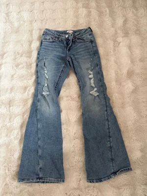 Blå bootcut low waist jeans från Gina Tricot - Säljer ett par blå bootcut jeans från Gina Tricot med slitningar på båda benen. Jeansen har klassisk femficksdesign, lila sömmar och är tillverkade i denim. Perfekt för dig som gillar en avslappnad och trendig look.