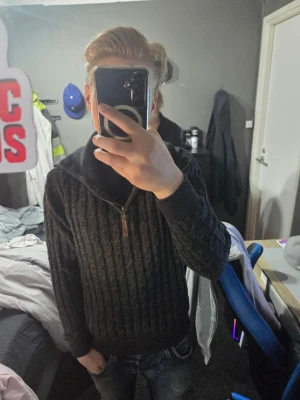 Mörkgrå stickad tröja med dragkedja - Snygg mörkgrå stickad tröja från Ragwear med halv dragkedja och hög krage. Tröjan har ett klassiskt kabelstickat mönster och ribbade muddar vid ärmslut och nederkant. Perfekt för lager-på-lager och passar till jeans eller chinos.