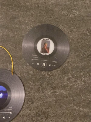 Dekorativa vinylskivor med albumomslag Frank ocean och daniel ceaser  - Två dekorativa vinylskivor i svart plast med tryckta albumomslag och låtinfo. Ena skivan har Frank Oceans 'Blond'-omslag, den andra har ett blått motiv och texten 'Cool' av Daniel Caesar. Perfekt som väggdekoration, båda har detaljer som liknar musikspelare och en har gul snöre för upphängning.
