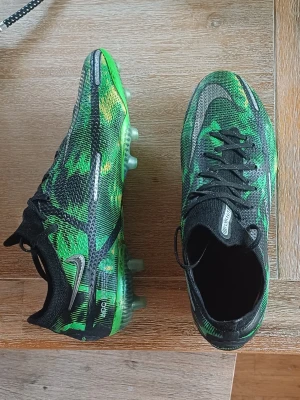 Nike Phantom GT2 Elite fotbollsskor - Säljer ett par Nike Phantom GT2 Elite fotbollsskor i grön, svart och gul färg. Skorna har en snygg mönstrad ovandel i syntetmaterial, snörning och dobbar för spel på konst gräs. Storlek 39.