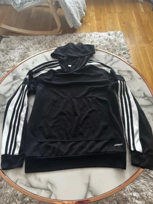 Svart Adidas hoodie med vita ränder - Svart hoodie från Adidas med klassiska vita ränder längs ärmarna och stor känguruficka framtill. Tillverkad i ett sportigt material och har Aeroready-tryck på ena sidan. Perfekt för en sportig och avslappnad stil.