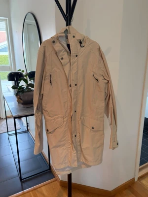 Beige parkas med huva och fickor - Lång beige parkas med justerbar huva och dragkedja framtill. Jackan har stora fickor med lock och knapp, samt justerbara ärmslut. Perfekt för kyliga dagar och enkel att matcha med olika outfits.