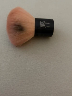 Kabukiborste från Livarno Home - Kabukiborste med täta, mjuka borst i ljusrosa med mörkare topp. Handtaget är svart och kompakt, perfekt för applicering av puder eller bronzer. Borsten har en rundad form som ger jämn finish och är enkel att använda för både ansikte och dekolletage.