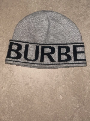 Grå Burberry mössa med logga - Snygg grå stickad mössa från Burberry med bred uppvikt kant och stor svart logotyp runt hela mössan. Klassisk och stilren design som passar perfekt till kalla dagar. Materialet är mjukt och bekvämt, perfekt för att hålla värmen.
