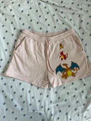 Beiga shorts med Pokémon-tryck - Snygga ljus rosa / peach / beiga shorts från Pokémon x asos design med tryck av Charmander, Charmeleon och Charizard på ena benet. Shortsen har resår och snörning i midjan samt fickor på sidorna och en bakficka. Perfekta för sommaren och chill dagar.