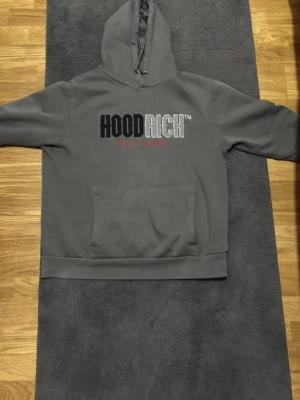 Grå hoodie från Hoodrich med tryck - Säljer en grå hoodie från Hoodrich med stor logga i svart och silver på bröstet samt röd text under. Hoodien har huva med dragsko och en stor magficka. Perfekt för dig som gillar streetwear och vill ha en snygg och bekväm look.
