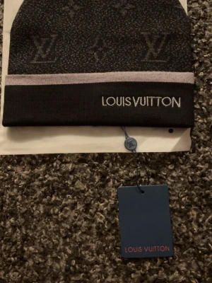 Svart mössa - Snygg svart mössa från med diskret-mönster och broderad logga framtill. Mössan har en grå kant och är stickad för extra komfort. Perfekt accessoar för att lyfta din stil under kyliga dagar.