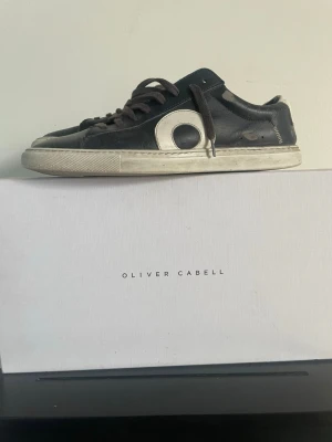 Svarta Oliver Cabell sneakers i läder - Snygga svarta sneakers från Oliver Cabell i läder med vit sula och vit detalj på sidan. Klassisk låg modell med svarta skosnören och diskret branding på hälen. Perfekta för dig som gillar stilrena och trendiga skor. Finns några slitningar utöver dom som ska vara där 