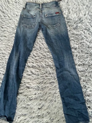 Blå slitna jeans med raka ben - Säljer ett par lågmidjade blå jeans från Replay med raka ben och slitningar på knäna. Jeansen har klassisk femficksdesign, låg midja och tydliga slitningar framtill för en cool, avslappnad look. Perfekta för dig som gillar streetstyle.