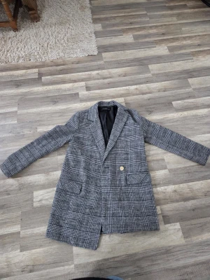 Grå rutig kappa från Zara Basic - Stilren grå kappa från Zara Basic med klassiskt rutigt mönster och svart innerfoder. Kappan har två framfickor, en knapp framtill och en rak passform som ger en snygg siluett. Perfekt för dig som gillar tidlös och trendig stil. Inköpt med fel storlek 