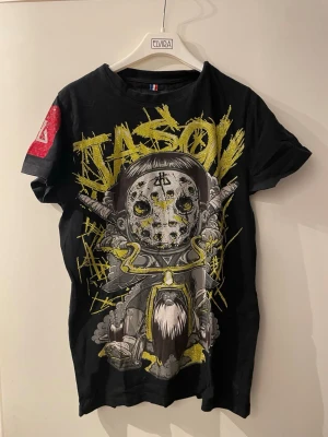 Horspist T-shirt - Cool svart t-shirt med stort grafiskt tryck av Jason och två svärd på framsidan. Texten 'JASON' i gult ovanför motivet och detaljer i grått, vitt och gult. Kortärmad modell med röd detalj på ena ärmen. Perfekt för dig som gillar streetwear och unika prints.