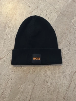 Svart mössa från BOSS - En stilren svart mössa från BOSS med uppvikt kant och en svart patch med orange BOSS-logga framtill. Mössan är ribbstickad och har en klassisk form som passar perfekt till streetwear och vardagsstil. Materialet är mjukt och bekvämt. Pris kan diskuteras. Ej använd