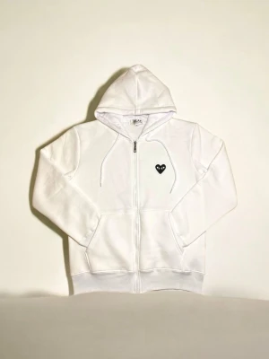 Vit zip hoodie från Comme des Garçons - Vit hoodie med dragkedja från Comme des Garçons Play. Klassisk passform med huva och dragsko, samt fickor framtill. På bröstet sitter det ikoniska svarta hjärta-ögon-logotypen. Perfekt för en clean och stilren look.