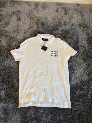 Vit Peak Performance piké t-shirt - Säljer en fräsch vit piké t-shirt från Peak Performance med svart logotyptryck på bröstet. Klassisk krage med knappar och korta ärmar. Perfekt till jeans eller shorts för en clean och sportig look.