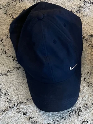 Mörkblå Nike keps med broderad logo - Snygg mörkblå keps från Nike med den klassiska broderade swoosh-loggan i vitt framtill. Kepsen har böjd skärm och justerbar rem bak. Tillverkad i ett mjukt och slitstarkt bomullsmaterial som passar perfekt till streetwear-stilen.