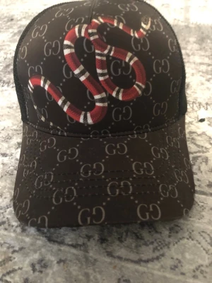 Gucci keps med orm och GG-mönster - A+ Först till kvarn går o scanna o allt A+ riktigt fett. Snygg keps från Gucci i svart  med klassiskt GG-mönster och en röd, vit och svart orm på framsidan. Kepsen har mesh på baksidan och justerbar passform. Tillverkad i Italien och perfekt för dig som gillar exklusiva accessoarer.