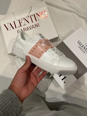 Valentino Garavani skor - Snygga vita sneakers från Valentino Garavani med bred rosa rem över vristen dekorerad med guldiga nitar. Skorna har en chunky sula och är tillverkade i slätt läder. Perfekta för dig som vill ha en lyxig och trendig look.