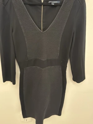 Svart kortklänning från French Connection - Svart kortklänning från French Connection med v-ringning och trekvartsärm. Klänningen har en strukturerad textil framtill och en diskret dragkedja i ryggen. Perfekt för dig som gillar stilrena och moderna plagg.