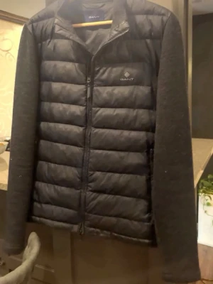 Marinblå  pufferjacka från GANT - Snygg marinblå cardigan från GANT med quiltad front och rygg samt mjuka stickade ärmar. Jackan har dragkedja framtill och diskret GANT-logga på bröstet. Perfekt för kalla dagar och ger en clean, modern look.