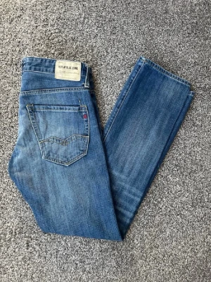 Blå raka jeans från Replay - Säljer ett par klassiska blå jeans från Replay med raka ben och snygga slitningar. Jeansen har fem fickor, tydliga sömmar och Replay Blue Jeans-märke bak i midjan. Tillverkade i bomull för en skön och avslappnad look.