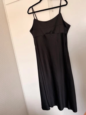 Svart långklänning med smala axelband - Stilren svart klänning med smala ställbara axelband och lätt glansigt tyg. Klänningen slutar vid smalbenen på mig som är 169 (längre midi-längd). Klänningen har en enkel, rak passform och har en dragkedja i sidan. Perfekt för dig som gillar minimalistisk och elegant stil. Superfin!! Obs djur finns i hemmet 