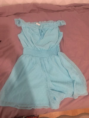 Ljusblå off shoulder jumpsuit - Supersöt ljusblå jumpsuit med off shoulder-modell och volangkant. Smockad midja för skön passform och små prickar i tyget som ger en extra detalj. Tunn och luftig, perfekt för sommardagar. Knytband framtill och korta ben.