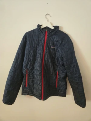 Mörkblå pufferjacka från Patagonia - Snygg mörkblå pufferjacka från Patagonia med röda dragkedjor och quiltat mönster. Jackan har hög krage, två sidofickor och är tillverkad i lätt, värmande material som passar perfekt för kalla vinterdagar.