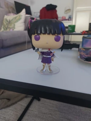 Funko Pop animefigur med lila outfit - Säljer en Funko Pop-figur föreställande en animekaraktär med svart hår, lila ögon och lila klänning. Figuren har vita detaljer, bälte och stövlar samt står på en genomskinlig plastbas. Perfekt till cosplay eller maskerad.