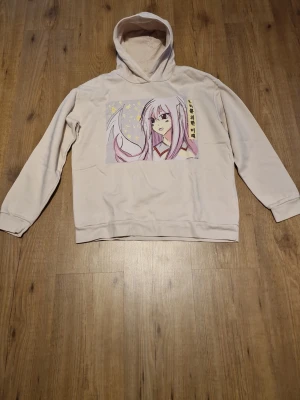 Beige hoodie  - Säljer en beige hoodie med huva och ett stort anime-tryck på bröstet i rosa och lila toner. Hoodien har långa ärmar, ribbade muddar och är gjord i mjukt bomullsliknande material. Perfekt för dig har en unik stil.