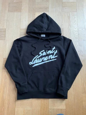 Svart Saint Laurent hoodie - Snygg svart hoodie från Saint Laurent (YSL) i storlek S med stor vit logga framtill. Klassisk modell med huva, dragsnören och stor magficka. Tillverkad i mjukt och bekvämt material som känns skönt mot huden. Hoodien är i mycket bra skick, utan hål, fläckar eller andra defekter. Äkthetsbevis finns. Perfekt för en avslappnad men stilren och trendig look. Hör gärna av dig vid frågor Mvh Olof!