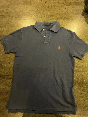 Royal blå pikétröja från Ralph Lauren - Klassisk blå pikétröja från Ralph Lauren med orange broderad logga på bröstet. Tröjan har korta ärmar, krage och två knappar framtill. Slim fit-modell som passar perfekt till jeans eller shorts.