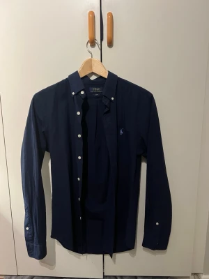 Mörkblå skjorta från Polo Ralph Lauren - Klassisk mörkblå skjorta från Polo Ralph Lauren med button-down krage och vita knappar. Skjortan har lång ärm och broderad logga på bröstet. Tillverkad i bomull för en bekväm och stilren look. Ej använd då den var för liten för mig