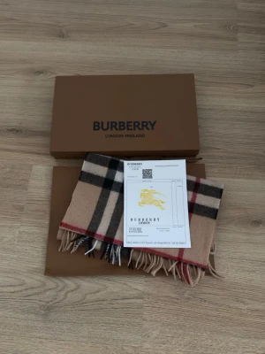 Beige rutig halsduk från Burberry - Klassisk beige halsduk från Burberry i 100% kashmir. Den har det ikoniska rutmönstret i svart, vitt och rött samt fransar i ändarna. Tillverkad i Skottland och levereras med originalkartong och certifikat.