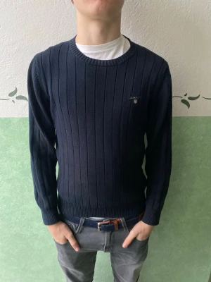 Mörkblå stickad tröja från GANT - Säljer en mörkblå stickad tröja från GANT med diskret logotyp på bröstet. Tröjan har ribbade muddar och rund hals, samt ett snyggt vertikalt mönster. Perfekt för dig som gillar klassisk och stilren design.