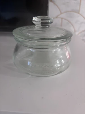 Rund glasburk med lock - Säljer en rund glasburk med tillhörande lock. Burken är helt genomskinlig och har en enkel, klassisk design med ett greppvänligt handtag på locket. Perfekt för förvaring av småsaker, godis eller kryddor.höjd 9cm diameter 11cm