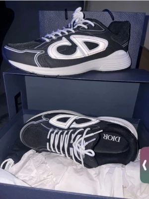 Svarta Dior sneakers med vita detaljer - Snygga svarta Dior sneakers med vita detaljer och stora CD-loggor på sidorna. Skorna har vita snören, tjock vit sula och mesh- samt läderpaneler för en modern look. Perfekta för dig som vill sticka ut med exklusiva och trendiga sneakers.