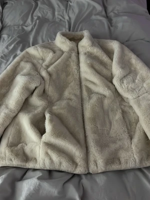 Beige teddyjacka med dragkedja - Mysig beige jacka med hög krage och dragkedja framtill. Jackan har en oversized passform och är tillverkad i mjukt, fluffigt material som ger en riktigt skön känsla. Perfekt för kyliga dagar när du vill vara både varm och stylish.