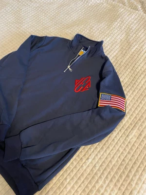Jeffrey Epstein Bomull Halfzip - Jeffrey Epsteins Quarter Zip USA flag med halfzip och ståkrage. På bröstet finns ett rött broderat märke och på ärmen sitter en patch med amerikanska flaggan. Tillverkad i mjukt bomullsmaterial och har ribbade muddar vid ärmslut och nederkant.