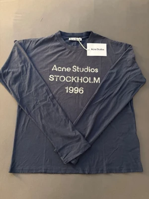 Mörkblå långärmad Acne Studios tröja - Snygg mörkblå långärmad tröja från Acne Studios med tryck 'Acne Studios STOCKHOLM 1996' på bröstet. Tillverkad i mjuk bomull med rund hals och avslappnad passform. Perfekt för en clean och stilren look.