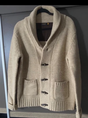 Beige stickad kofta med träknappar - Säljer en beige stickad kofta från Review med grov ribbad krage och fyra träknappar framtill. Koftan har två stora fickor och ribbade muddar vid ärmslut och nederkant. Perfekt för lager på lager och en avslappnad stil. En knapp har ramlat av, men finns kvar om man vill sy på den