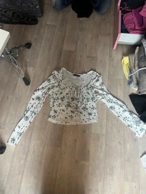 Blommig långärmad blus från SHEIN - Söt blus från SHEIN i vitt med småblommigt mönster i blått och gul. Blusen har långa ärmar, fyrkantig ringning och en dekorativ knytning framtill. Tillverkad i ett ribbat, stretchigt material som sitter bekvämt.