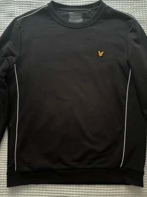 Svart sweter tröja från Lyle & Scott - Svart sweter tröja från Lyle & Scott med rund halsringning och vita detaljer längs sidorna och axlarna. Tröjan har den klassiska gula logon broderad på bröstet och ribbade muddar vid ärmslut och nederkant. Perfekt för en stilren och enkel look.
