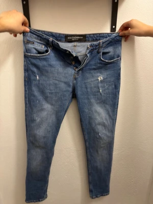 Blå jeans från Dolce & Gabbana - Säljer ett par blå jeans från Dolce & Gabbana med knäppning fram och slitna detaljer på låret. Klassisk femficksmodell med patch på bakfickan. Jeansen har en smal passform och är tillverkade i denim.